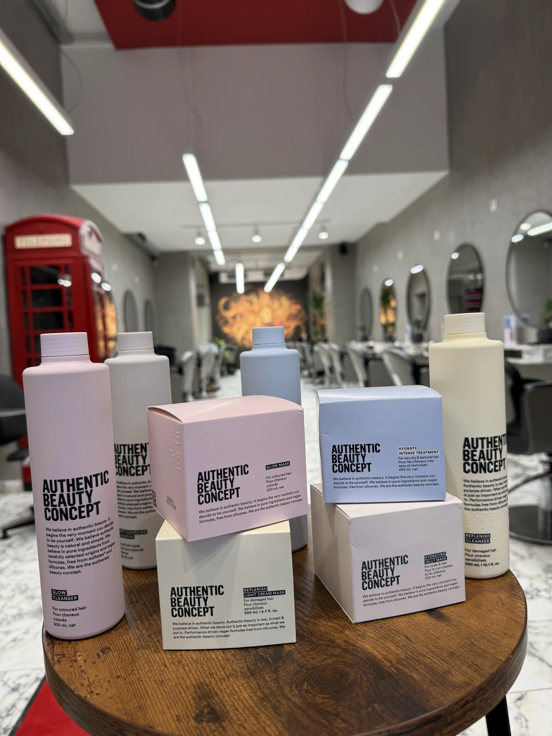 Hairfashion by Edmond - Haarschnitt und Styling - Nahaufnahme von großen Flaschen und Schachteln mit Haarpflegeprodukten von Authentic Beauty Concept auf einem runden Holztisch in einem Salonambiente; im Hintergrund ist eine rote Telefonzelle zu sehen.