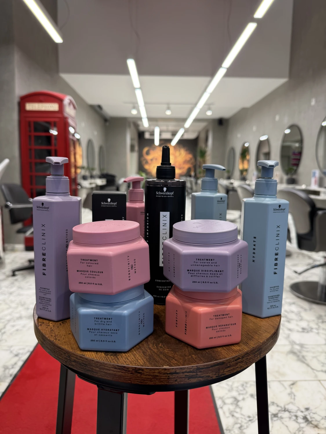 Hairfashion by Edmond - Haarschnitt und Styling - Eine Auswahl an Haarpflegeprodukten in Rosa, Blau und Schwarz aus den Produktlinien Fibre Clinix und Clinix ist auf einem kleinen Holztisch in einem Friseursalon ausgestellt; im Hintergrund ist eine rote Telefonzelle zu sehen.