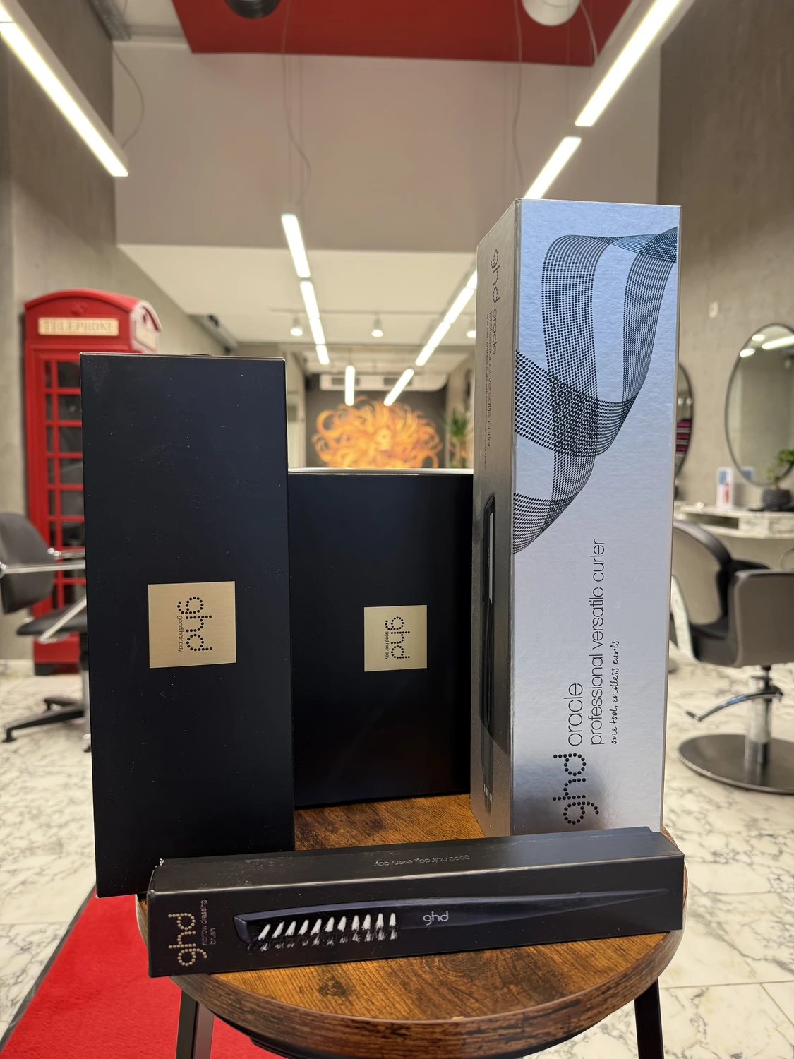 Hairfashion by Edmond - Haarschnitt und Styling -Mehrere schwarze und silberne ghd-Haarpflegeproduktverpackungen, darunter ghd Oracle und ein ghd Styler, sind auf einem Holztisch in einem Friseursalon ausgestellt.